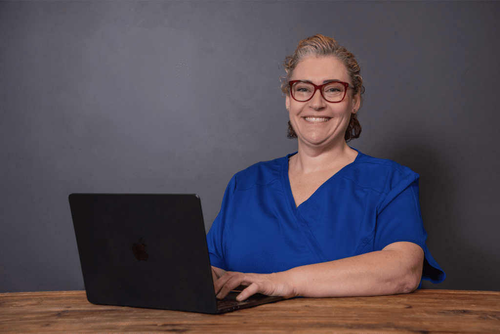 Woman in blue scrubs smiling at desk with laptop. Medical professional headshot Brisbane style with clean, neutral backdrop.