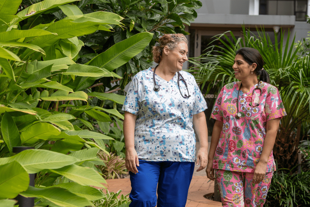 Two nurses walking through lush garden, laughing in scrubs with stethoscopes. Warm, friendly team image by professional headshot Brisbane expert.